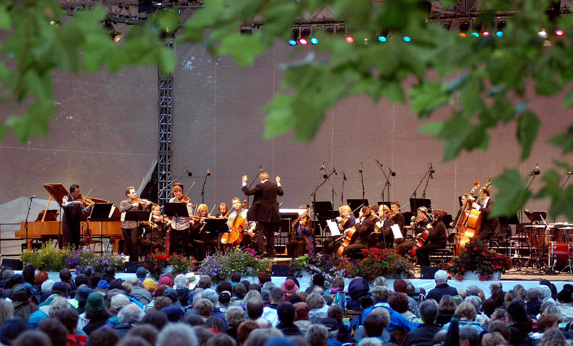 Blick durch Blätter hindurch auf eine Bühne mit einem klassischen Orchester. Davor das Publikum