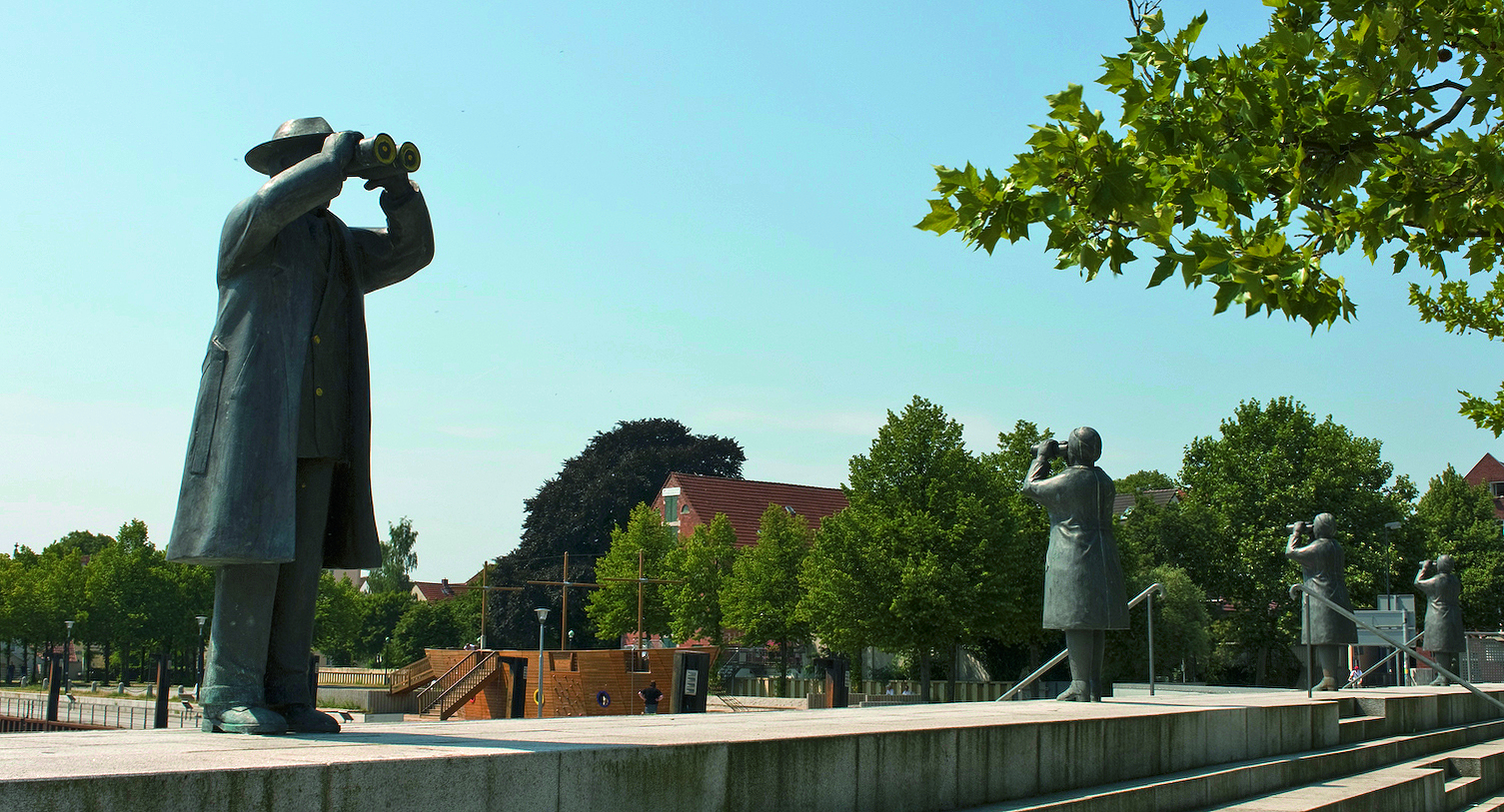 Statue zeigt Männer mit Ferngals am Rande eines Hafenbeckens mit Segelbooten