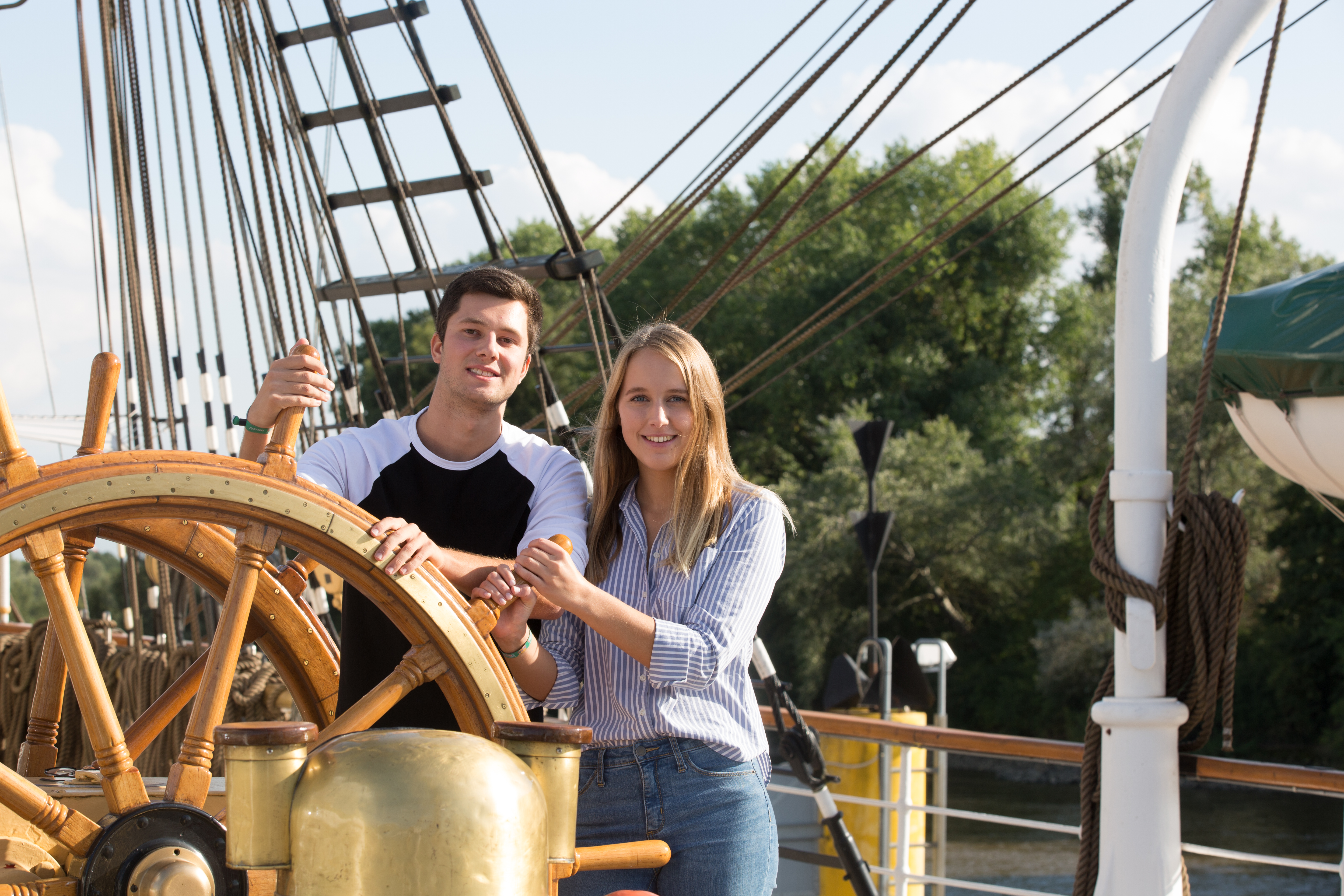 Junges Paar steht am Ruder eines Schiffs und schaut in die Kamera