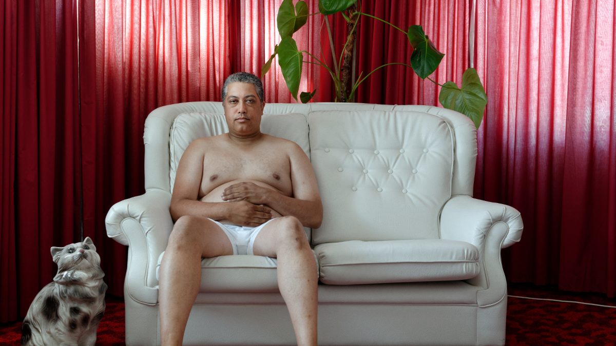 Mann sitzt in Unterhose aus hellem Sofa und hält sich die Hände vor den Bauch