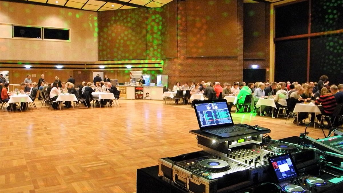 Ein DJ-Pult und viele an Tischen essende Menschen in einer großen Halle