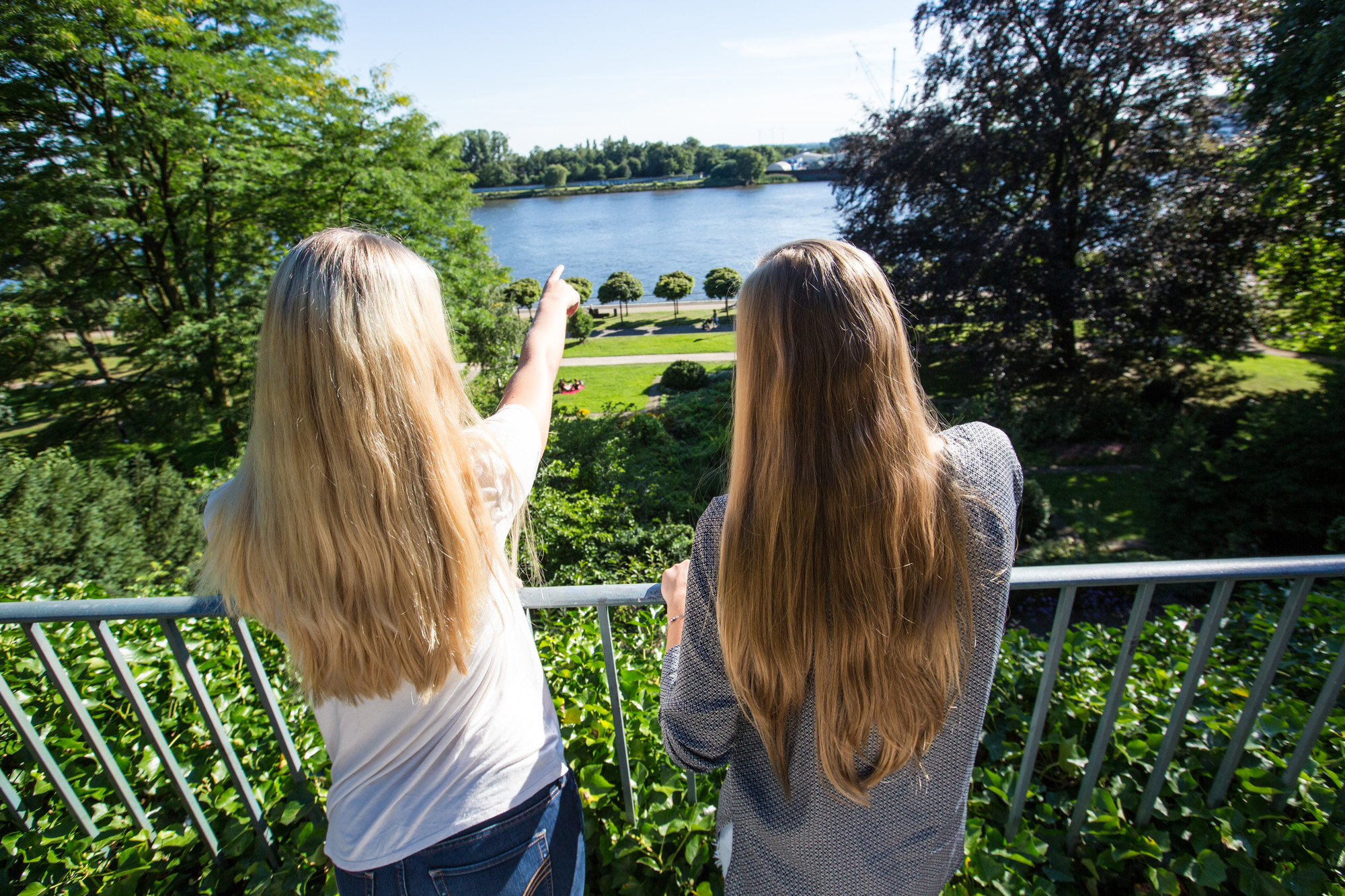 Zwei Frauen stehen am Geländer in einem Park und schauen in die Ferne. Die linke Frau zeigt mit dem Finger nach vorne.