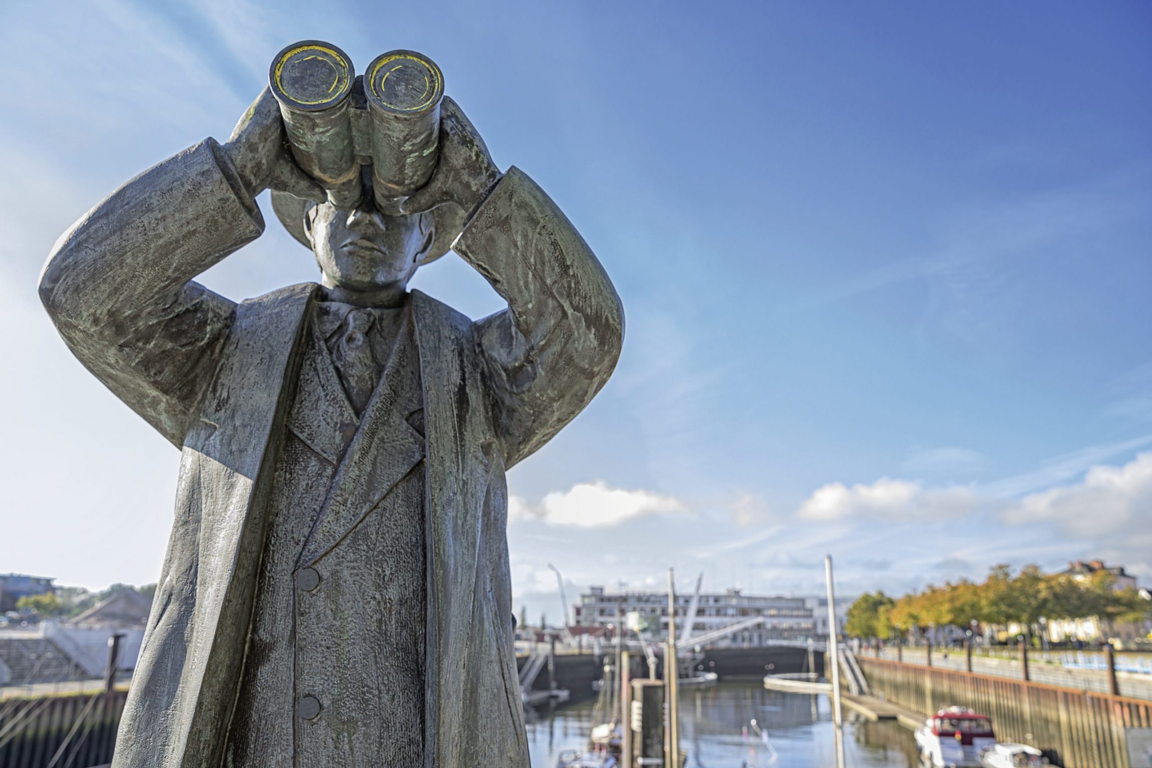 Statue von einem Mann, der durch ein Fernglas schaut. Im Hintergrund sieht man den Vegesacker Hafen.