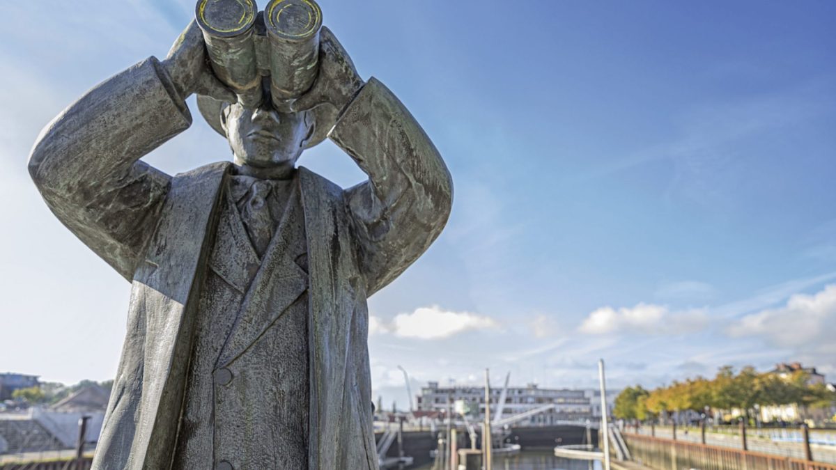 Statue von einem Mann, der durch ein Fernglas schaut. Im Hintergrund sieht man den Vegesacker Hafen.