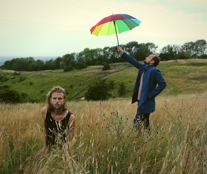 Zwei Männer sind in einem Kornfeld. Einer hält einen bunten Regenschirm in die Luft