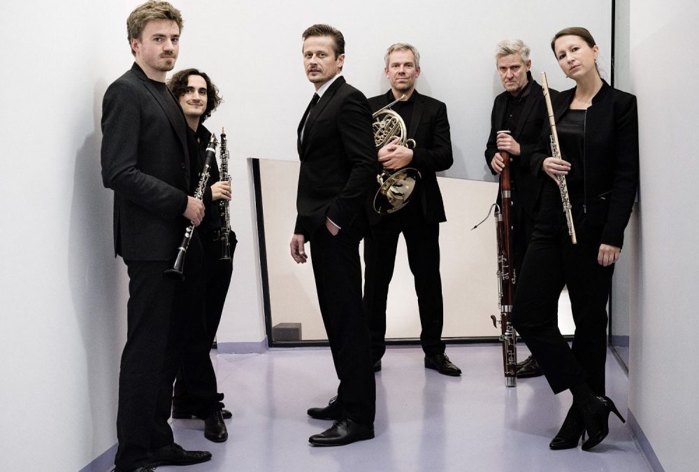 6 Personen in schwarzen Anzügen stehen in einem weißen Raum und halten verschiedene Musikinstrumente in der Hand
