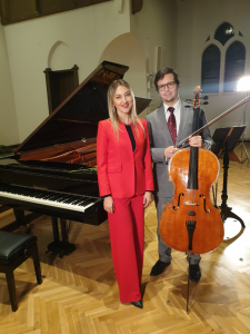 Eine Frau in einem roten Anzug und ein Mann in einem gruaen Anzug stehen vor einem Flügel. Der Mann hält außerdem ein Cello in der Hand