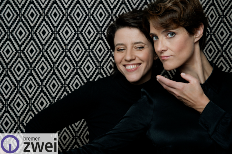 Zwei Frauen stehen lachend vor einer schwarzweiß gemusterten Wand.