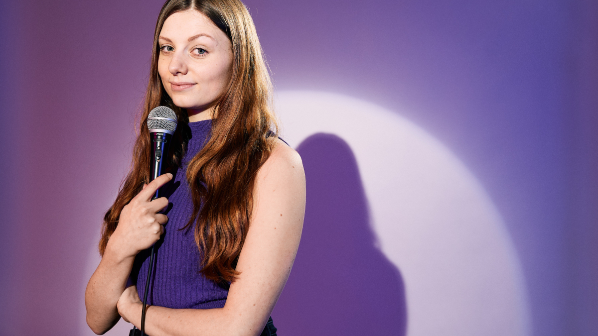 Lara Ermer steht vor einem lila Hintergrund. In der Hand hält sie vor ihrem Mund ein Mikrofon.