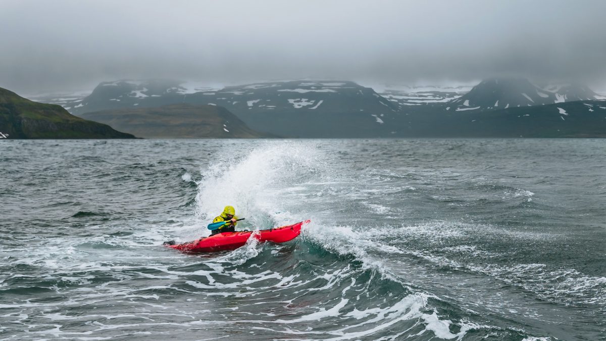 Ein Mann paddelt mit dem Kajak durch das unruhige Meer. Im Hintergrund sind Berge zu erkennen.