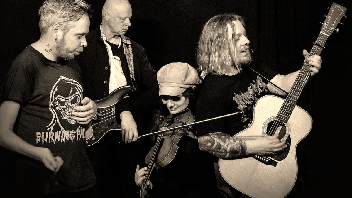 Schwarzweiß Bild von vier Männern, die nebeneinander stehen und sitzen und ihre Instrumente spielen