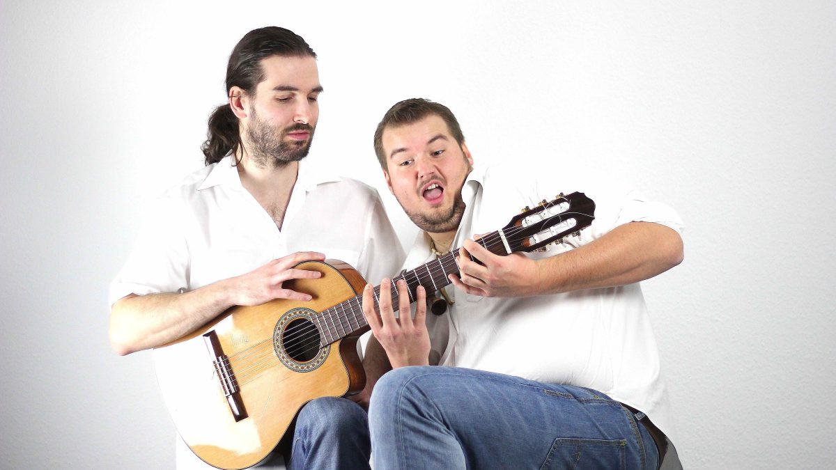 Zwei Männer in weißen Hemnden und blauen Jeans sitzen nebeneinander und versuchen aus Spaß zu zweit eine Akustikgitarre zu spielen.