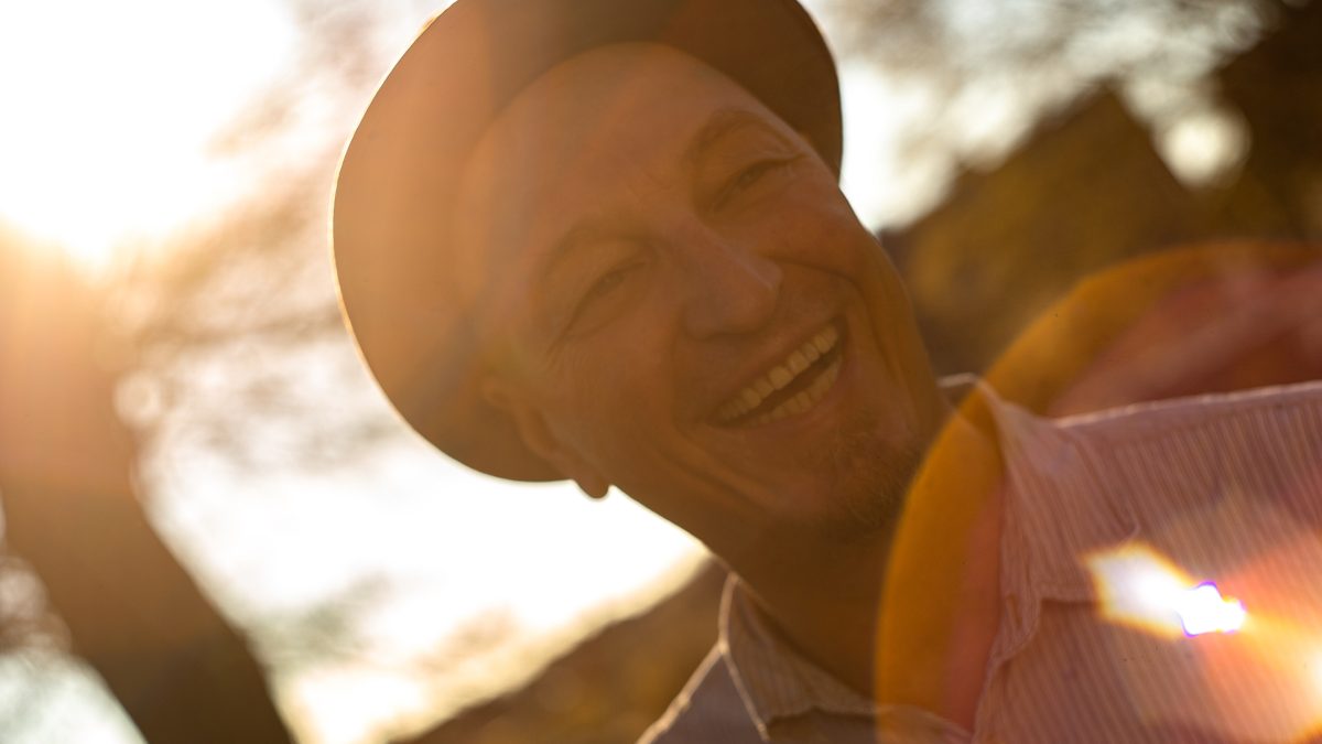 Portrait von einem lachendem Mann mit Hut. Die Sonne scheint von hinten auf ihn und in die Kamera