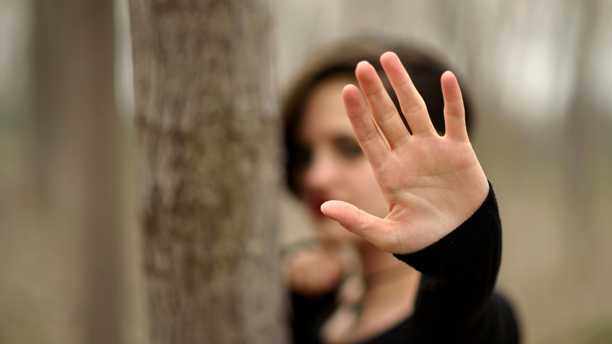Eine Frau an einem Baum streckt ihre flache Hand in die Kamera und symbolisiert Stopp.