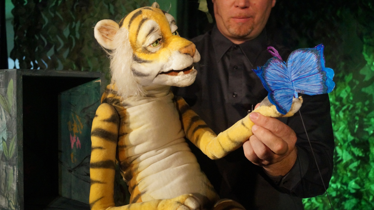 Eine große Figur eines Tigers sitzt neben einem kostümierten Mann auf einer Bühne.