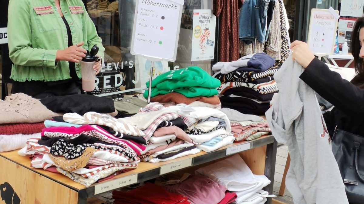 Eine Frau steht hinter einem Tisch. Auf dem Tisch leigen viele Kleidungsstücke, die zum Verkauf angeboten werden. Vor dem Tisch lässt sich eine weitere Frau erkennen, die sich ein Kleidungsstück genauer ansieht.