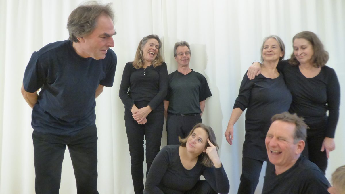 Eine Gruppen Menschen, alle in schwarz gekleidet, steht vor einem weißen Hintergrund zusammen und lachen