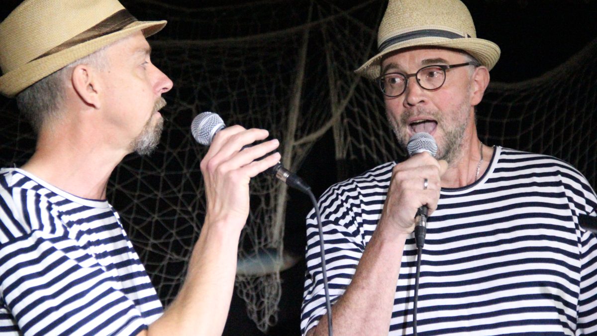 Zwei Männer in gestreiften T-Shirts und mit Hüten stehen nebeneinander und singen zusammen
