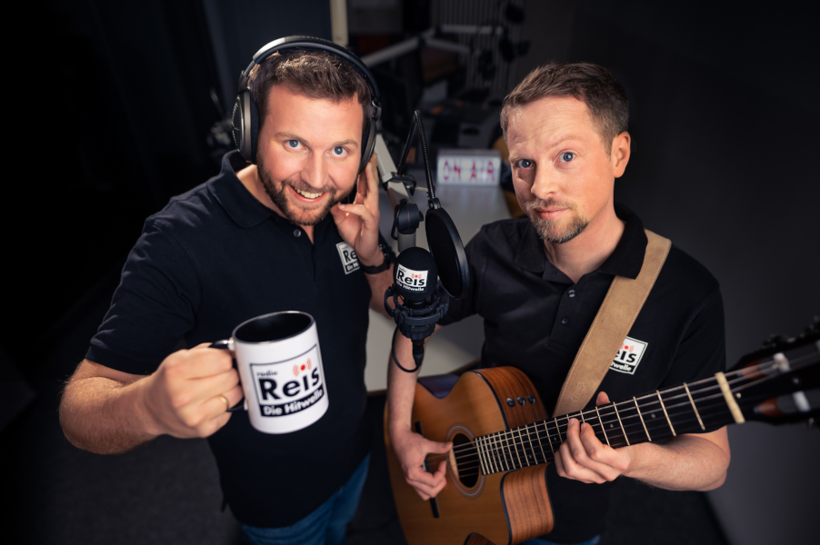 Zwei Männer stehen in einem Radiostudio. Einer hält eine Tasse in der Hand, der andere spielt Gitarre