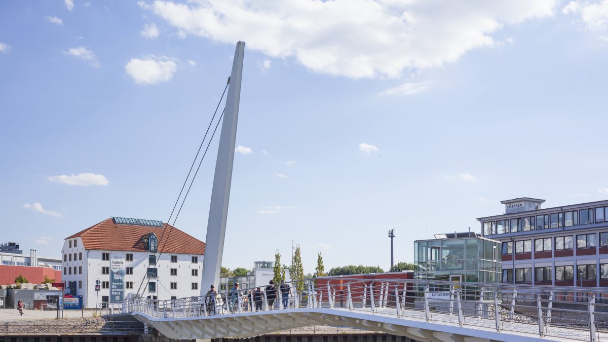 Eine weiße Brücke führt über den Vegesacker Museumshaven. Auf der Brücken schieben zwei Menschen ihre Fahrräder. Im Hintergrund ist das Vegesacker Geschichtenhaus zu erkennen.