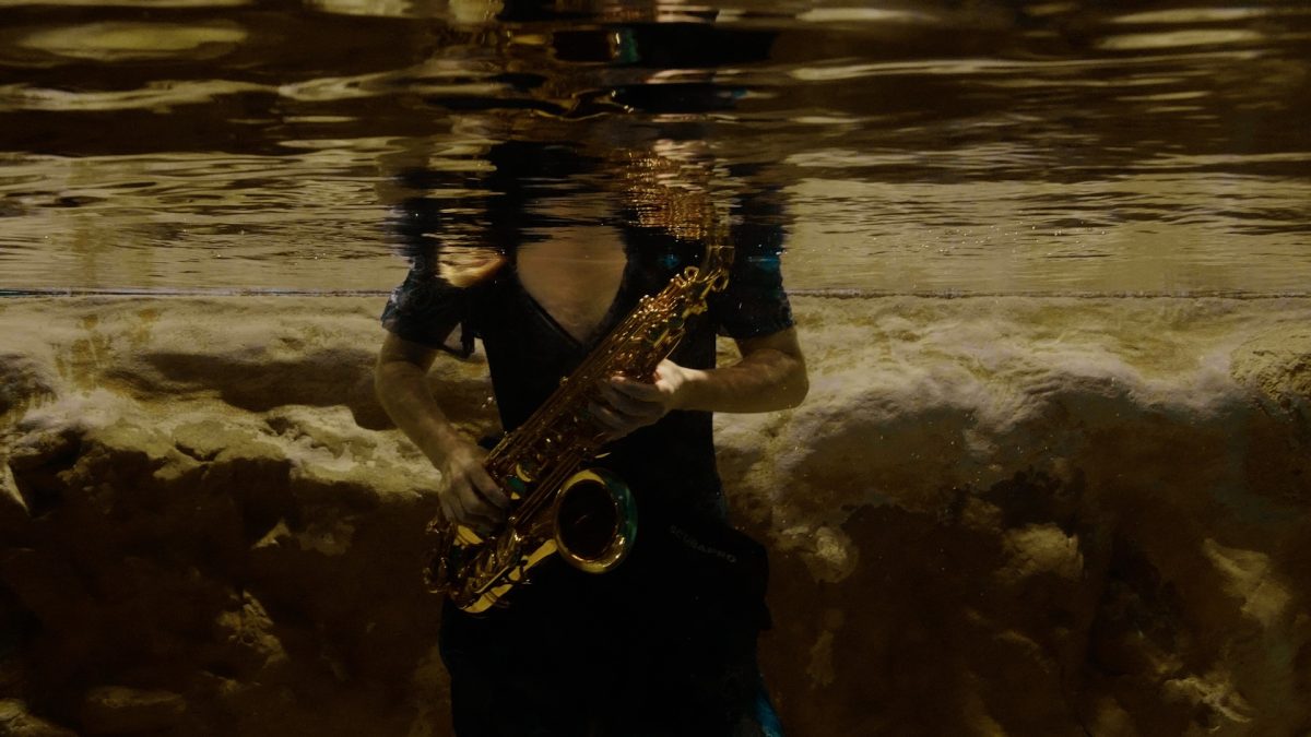 Unterwasser Foto von einer Frau in einem blauen Kleid, die Saxofon spielt. Der Kopf ist nicht zu sehen.