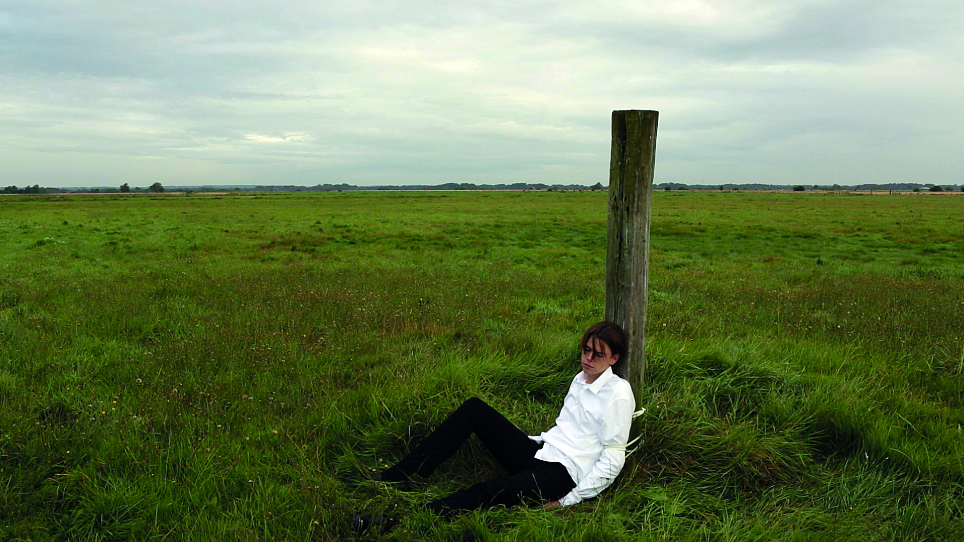 Ein junger Mann im weißen Hemd und schwarzer Hose sitzt mit dem Rücken angebunden an einem Holzpfahl. Um ihn herum sind grüne Wiesen.