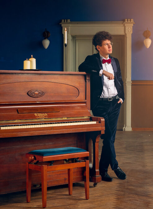 Ein junger Mann im Anzug lehnt an ein braunes Piano