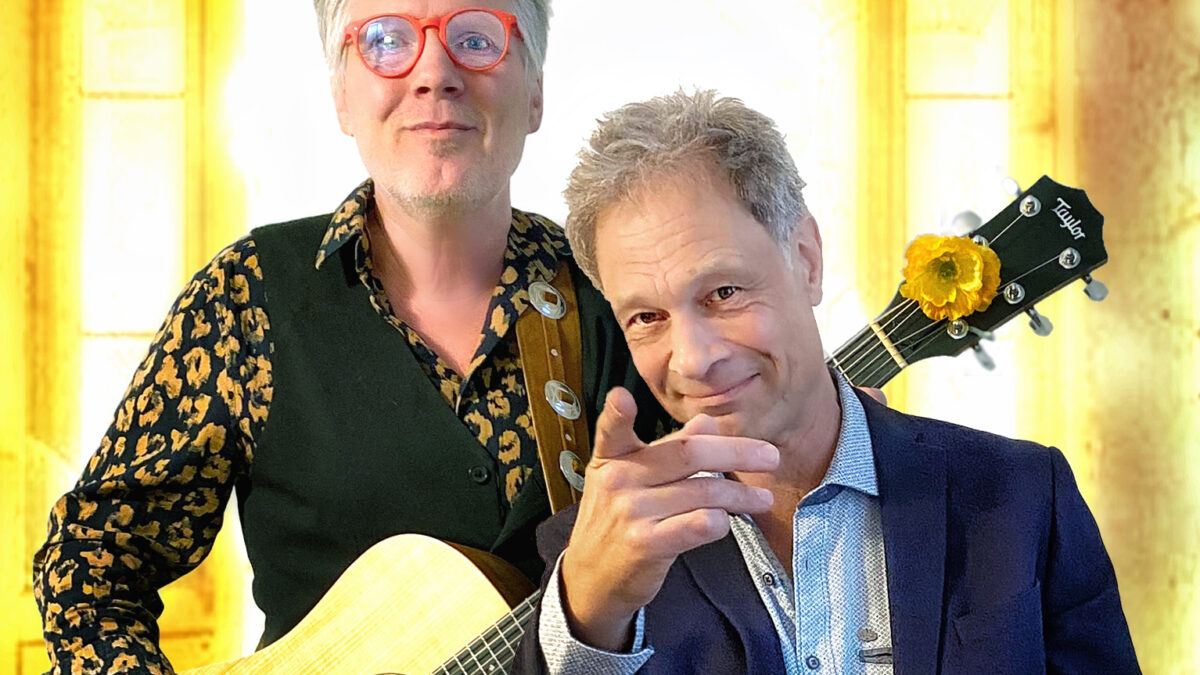 Jürgen Schöffel mit Gitarre leicht versetzt hinter Martin Heckmann, der mit Mikrofon auf einem Stuhl sitzt und in die Kamera zeigt